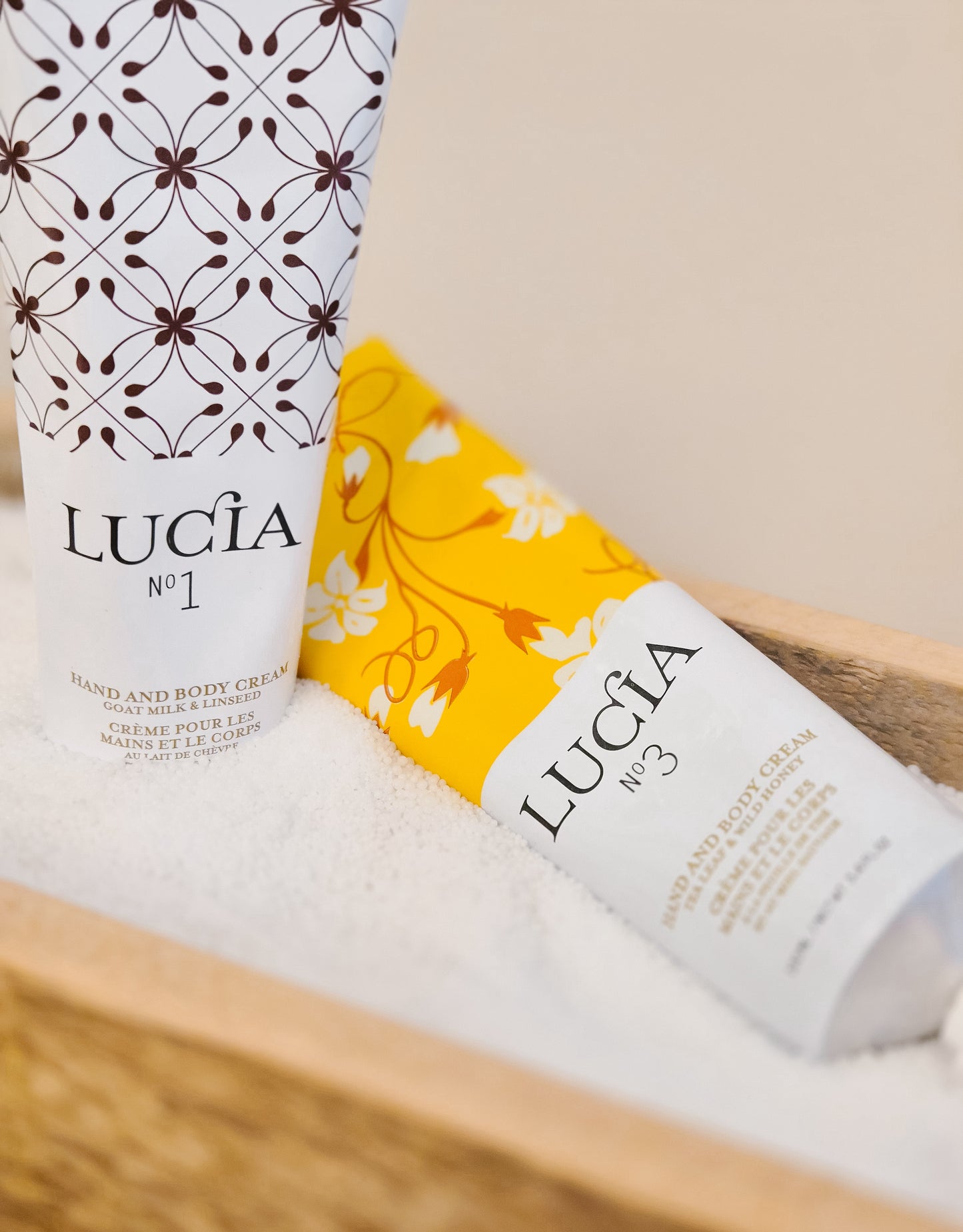 Two bottles of Lucia hand and body cream on a wooden surface with a neutral background.