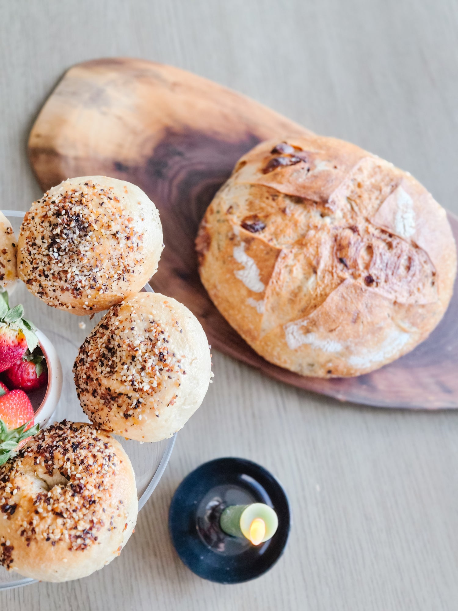Assorted bread loaves on a wooden cutting board with a bowl of strawberries and a candle.